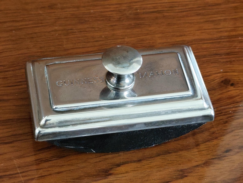Puede incluir: Un secante de tinta plateado con las palabras "GUINNESS MAHON" grabadas. Tiene un asa redondeada y una base rectangular con una almohadilla negra. El art&iacute;culo est&aacute; sobre una superficie de madera. Mide unos 10 cm de largo.