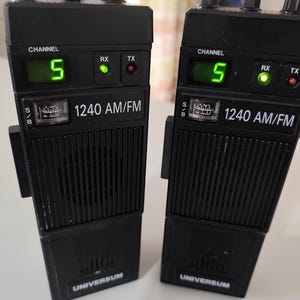Könnte beinhalten: Zwei schwarze Universum 1240 AM/FM Funkgeräte mit ausziehbaren Antennen. Jedes Gerät verfügt über eine digitale Kanalanzeige mit der Ziffer 5, ein analoges Signalmessgerät sowie RX- und TX-Anzeigeleuchten. Die Geräte haben ein Lautsprechergitter.