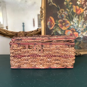 May include: A woven wicker basket with a red and brown pattern. The basket is closed and has a hinged lid. The basket is sitting on a dark green surface.