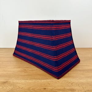 May include: A lampshade with a trapezoidal shape. The lampshade is navy blue with horizontal red stripes. The lampshade is sitting on a light brown wooden surface, with a white wall in the background.