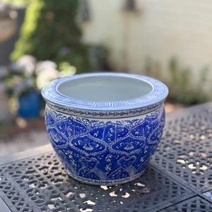 May include: A round ceramic planter featuring a deep blue and white intricate pattern. The rim is decorated with a geometric Greek key design and a triangular border. The body displays a repeating floral-inspired motif in white on blue.