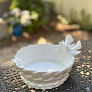 May include: A white ceramic bowl featuring a woven basket-weave texture on the exterior. The rim is decorated with a small sculpted flower on one side and two sculpted birds in flight on the opposite side, creating a detailed decorative accent.