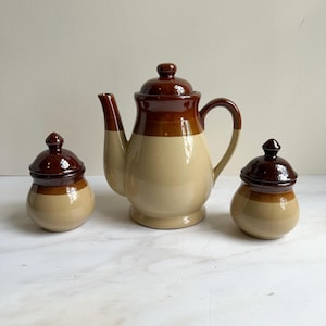 May include: A vintage ceramic tea set featuring a teapot and two lidded jars. The set has a two-tone design with a dark brown top and a tan base. The teapot has a curved spout and handle.