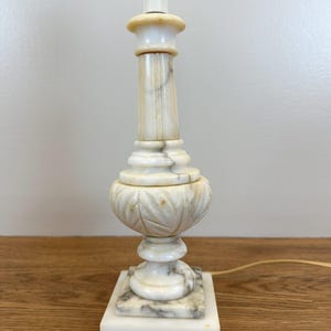 May include: A table lamp base crafted from white and grey veined marble. The structure features a square base, a central column with vertical fluting, and a rounded mid-section decorated with carved leaf patterns. A light-colored power cord extends from the back.