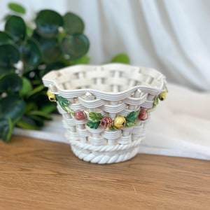 May include: A white ceramic basket with a woven texture and a scalloped rim. The exterior is decorated with a garland of small, colorful sculpted fruits and green leaves, including yellow and reddish-brown berries, encircling the middle section.