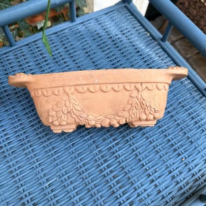 May include: A rectangular terracotta planter with rounded handles on each side. The exterior features a decorative relief pattern consisting of a scalloped border along the top edge and draped floral swags across the front, supported by four feet.