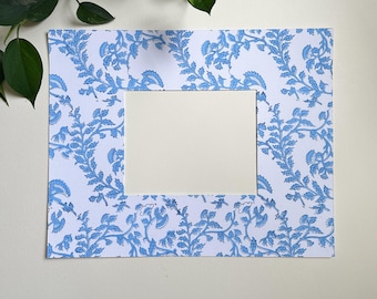 May include: A rectangular picture frame with a white center and a blue floral pattern border. The frame is made of paper and is placed against a cream-colored background. Green leaves are visible in the upper left corner.