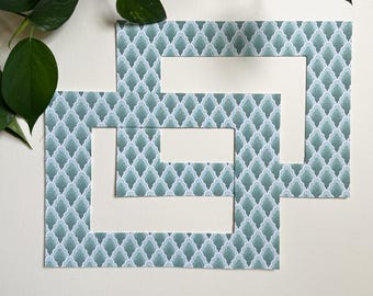 May include: Two decorative picture frames with a teal and white geometric pattern. The frames are cut out in a unique shape, with a white interior. Green leaves are visible in the background.