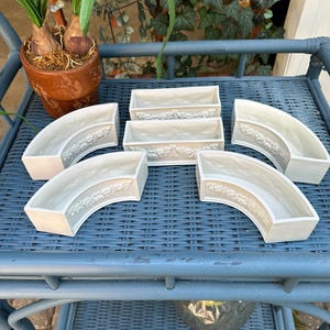 May include: A set of five off-white ceramic planters featuring two rectangular units and three curved units. Each planter has a raised floral relief pattern along the front exterior surface, providing a decorative texture to the smooth finish.