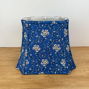 May include: A blue lampshade with a floral pattern. The fabric shade features white flowers and green vines against a blue background. The lampshade has a rectangular shape with a flared bottom and a white interior.