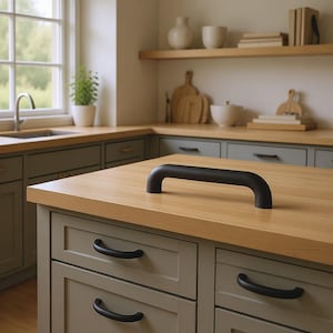 May include: A black, curved cabinet handle sits on a light wood worktop. The handle is a solid, rounded bar. The kitchen features grey cabinets, a window, and decorative items on shelves, suggesting a modern farmhouse style.