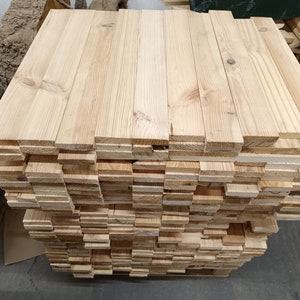 May include: A pile of untreated wooden planks, pale in colour. The rectangular boards are likely softwood, displaying visible grain and knots. The planks are neatly stacked, revealing both the end and side grain of the wood.