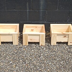 May include: Three wooden planter boxes, each with a square shape and a simple design. The boxes are made of natural wood and have a light brown colour.