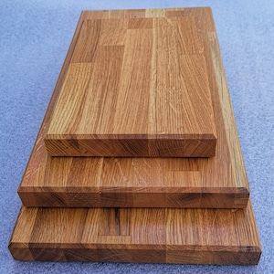 May include: Three wooden chopping boards stacked on top of each other. The boards are made of light brown wood with a natural grain pattern. The top board is the largest, followed by the middle board, and then the smallest board at the bottom.