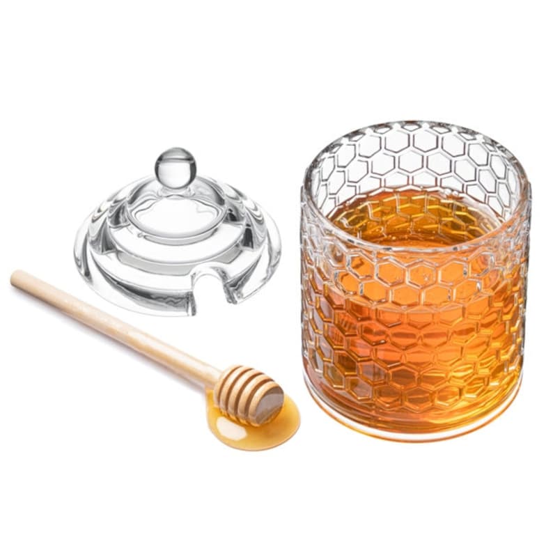 K&ouml;nnte beinhalten: Ein klares Glas Honigglas mit einem Wabenmuster, gef&uuml;llt mit Honig. Das Glas hat einen klaren Glasdeckel mit einem kleinen Knopf oben. Ein Holzhonigr&uuml;hrer liegt auf einer wei&szlig;en Oberfl&auml;che mit einer kleinen Honigpf&uuml;tze.