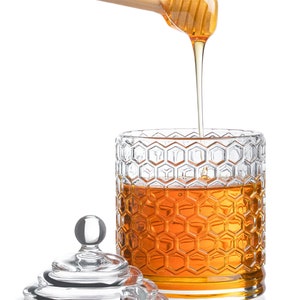 K&ouml;nnte beinhalten: Ein gl&auml;sernes Honigglas mit einem Wabenmuster und einem h&ouml;lzernen Honigl&ouml;ffel, der Honig in das Glas tropft. Das Glas hat einen durchsichtigen Glasdeckel.