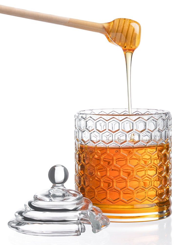 K&ouml;nnte beinhalten: Ein gl&auml;sernes Honigglas mit einem Wabenmuster und einem h&ouml;lzernen Honigl&ouml;ffel, der Honig in das Glas tropft. Das Glas hat einen durchsichtigen Glasdeckel.