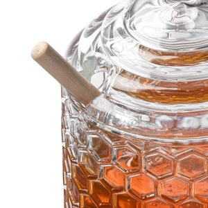 K&ouml;nnte beinhalten: Ein klares Glas Honigglas mit einem Wabenmuster und einem h&ouml;lzernen Honigl&ouml;ffel. Das Glas hat einen Deckel und ist mit Honig gef&uuml;llt.