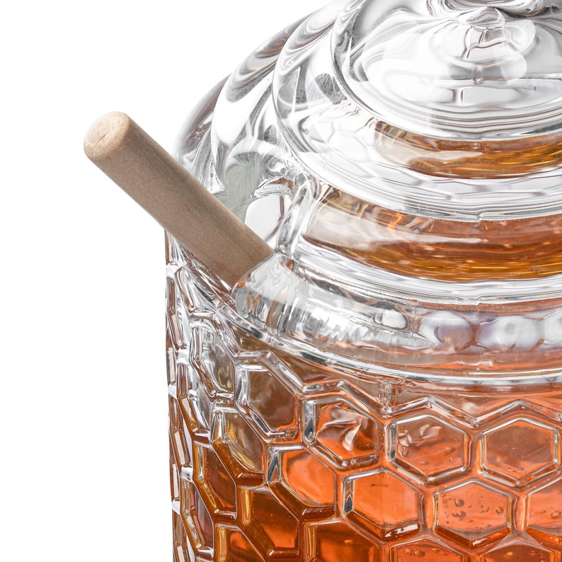 K&ouml;nnte beinhalten: Ein klares Glas Honigglas mit einem Wabenmuster und einem h&ouml;lzernen Honigl&ouml;ffel. Das Glas hat einen Deckel und ist mit Honig gef&uuml;llt.