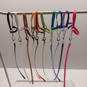 Könnte beinhalten: Eine Auswahl an farbigen Satinband-Lanyards mit Metallverschlüssen und Ketten. Die Lanyards sind in verschiedenen Farben erhältlich, darunter Schwarz, Weiß, Gold, Rot, Grün, Türkis, Lila und Pink. Sie hängen an einer weißen Metallstange.