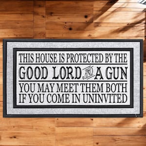 May include: Rectangular doormat with a black border and a gray inner border. The white center features black text: "THIS HOUSE IS PROTECTED BY THE GOOD LORD AND A GUN. YOU MAY MEET THEM BOTH IF YOU COME IN UNINVITED."