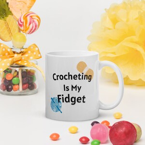 May include: White ceramic mug with the text "Crocheting Is My Fidget" in black, with a yarn ball and crochet hook graphic. The mug is surrounded by colorful candies and a jar of sweets.