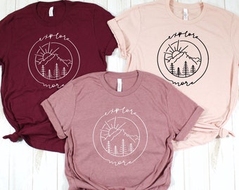 May include: Three t-shirts in burgundy, mauve, and light pink. Each shirt features a white circular design with a mountain, sun, and tree graphic, and the words "explore more". The t-shirts have rolled sleeves and a casual style.
