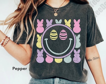 Pode incluir: Uma camiseta cinza com um gráfico desbotado de um rosto sorridente com ovos de Páscoa como olhos e doces Peeps em tons pastel que cercam o rosto. O texto "Pepper" é visível no canto inferior esquerdo da imagem.