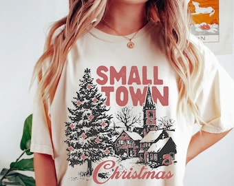 May include: Cream-colored t-shirt with a graphic featuring a Christmas tree, houses, and the words "SMALL TOWN Christmas" in red. The design evokes a cozy, festive holiday theme.