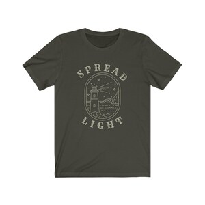 Op de afbeelding: Een donkergroen T-shirt met een witte omtrek van een vuurtoren, golven en sterren. De tekst "SPREAD LIGHT" staat boven en onder de afbeelding.