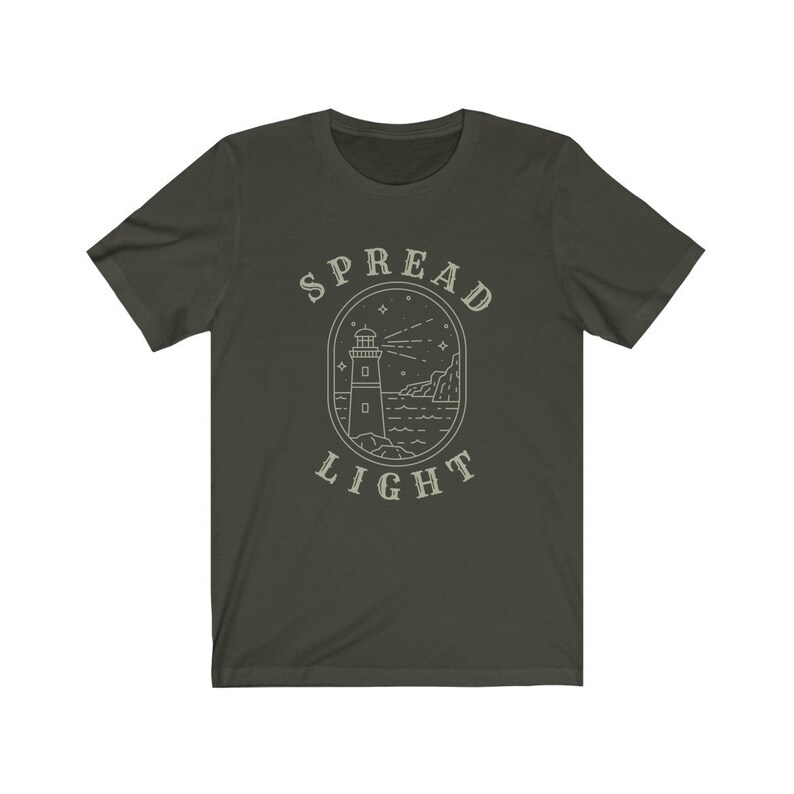 Op de afbeelding: Een donkergroen T-shirt met een witte omtrek van een vuurtoren, golven en sterren. De tekst "SPREAD LIGHT" staat boven en onder de afbeelding.