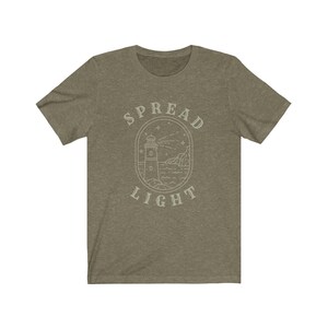 Op de afbeelding: Een groen T-shirt met een witte omtrek van een vuurtoren en de tekst "SPREAD LIGHT" in een cirkel eromheen.