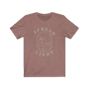 Op de afbeelding: Een heidekleurig t-shirt met een lichtgroen ontwerp van een vuurtoren met de tekst "SPREAD LIGHT" eromheen.