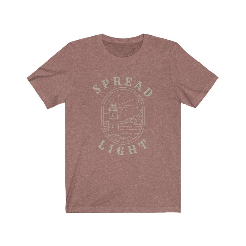 Op de afbeelding: Een heidekleurig t-shirt met een lichtgroen ontwerp van een vuurtoren met de tekst "SPREAD LIGHT" eromheen.