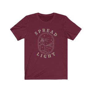 Op de afbeelding: Bordeauxkleurig T-shirt met een witte omtrek van een vuurtoren, golven en sterren. De tekst "SPREAD LIGHT" staat boven de vuurtoren.