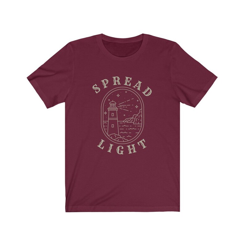 Op de afbeelding: Bordeauxkleurig T-shirt met een witte omtrek van een vuurtoren, golven en sterren. De tekst "SPREAD LIGHT" staat boven de vuurtoren.