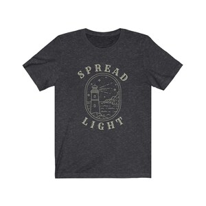 Op de afbeelding: Donkergrijs t-shirt met een vuurtoren afbeelding en de tekst "SPREAD LIGHT" in een witte omtrek.