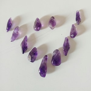 Puede incluir: Una colección de piedras preciosas de amatista, cada una con una forma única y diferentes tonos de púrpura. Las piedras están dispuestas sobre una superficie blanca, mostrando su apariencia natural y pulida. Estas piedras son ideales para la fabricación de joyas o la curación con cristales.