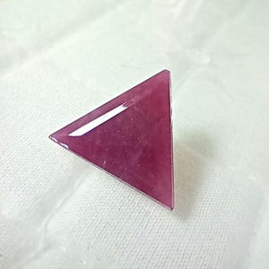 Op de afbeelding: Een driehoekige edelsteen met een diepe magenta-rode tint en een glad, gepolijst oppervlak. De steen heeft scherpe, strakke randen en een platte bovenkant, met een subtiele reflecterende glans over de gefacetteerde, geometrische vorm.