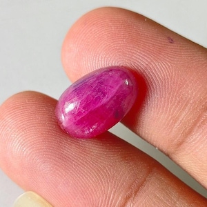 Pode incluir: Uma pequena pedra preciosa polida de formato oval com uma cor magenta-rosa vibrante e profunda. A pedra apresenta uma superfície lisa e arredondada com inclusões internas visíveis e textura natural, translúcida sob a luz.