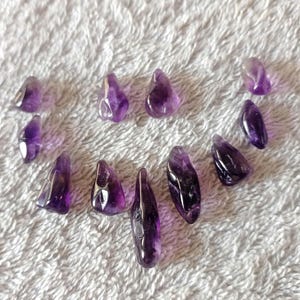 Puede incluir: Una colección de piedras preciosas de amatista pulidas de varias formas y tamaños. Las piedras muestran una gama de tonos púrpuras, desde lavanda claro hasta violeta oscuro, con áreas translúcidas y opacas. Las piedras preciosas están dispuestas sobre una superficie blanca texturizada.