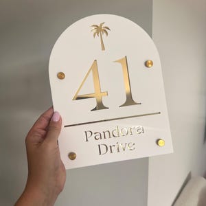 Puede incluir: Un letrero de dirección blanco con detalles dorados. El letrero muestra el número "41" en dorado, una palmera dorada y las palabras "Pandora Drive". Remaches dorados aseguran el letrero.