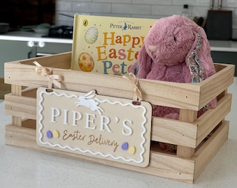May include: A wooden crate containing a Peter Rabbit book, a pink plush bunny, and a sign reading "Piper's Easter Delivery." The sign has a white border and a bunny cutout, perfect for Easter.