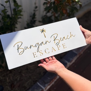 May include: White rectangular wooden sign with gold lettering that reads "Bungan Beach ESCAPE". A gold palm tree graphic is above the word "Bungan". The sign is held up by a person.
