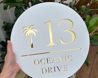 Puede incluir: Un letrero blanco y redondo con el número de la casa y detalles dorados. El número "13" se muestra de forma destacada, junto con una silueta de palmera y las palabras "OCEANIC DRIVE". El letrero se sostiene frente a un exuberante follaje verde.