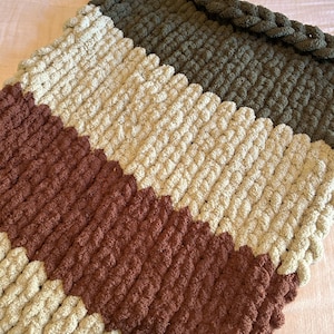 May include: A handmade, chunky knit blanket in a diagonal striped pattern. The blanket features alternating stripes of brown, cream, and olive green. The edge is finished with a braided trim. This cozy blanket is perfect for snuggling.