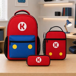 Könnte beinhalten: Ein roter und blauer Rucksack, eine passende Lunchtasche und ein Federmäppchen. Jeder Artikel zeigt einen weißen Kreis mit einem roten Buchstaben K in der Mitte. Rucksack und Lunchtasche haben zwei gelbe Kreise auf der blauen Vordertasche.