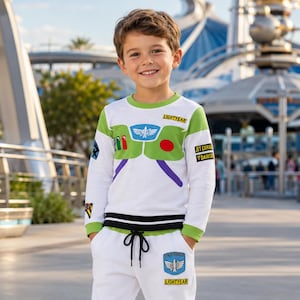 Puede incluir: Un conjunto de sudadera blanca de manga larga y pantalones deportivos a juego con detalles en verde y gráficos inspirados en un traje de guardián espacial. La parte superior incluye un emblema en el pecho y el pantalón tiene cordón.