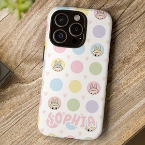 Pode incluir: Uma capa de telefone branca com um padrão de bolinhas coloridas e o nome "Sophia" em escrita rosa. A capa apresenta ilustrações de personagens de desenhos animados e círculos em tons pastel. A capa do telefone está em uma superfície de madeira.