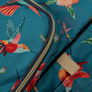 Peut inclure: Sac de couleur sarcelle avec un motif vibrant de colibris et de fleurs. Le sac est dot&eacute; d'une fermeture &eacute;clair dor&eacute;e et d'une sangle beige. Le motif comprend des accents rouges, oranges et verts, cr&eacute;ant une esth&eacute;tique color&eacute;e et attrayante.
